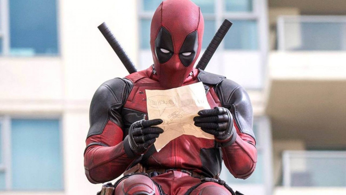 Deadpool dalam kostum merah dan hitam, sedang duduk sambil membaca tulisan di selembar kertas